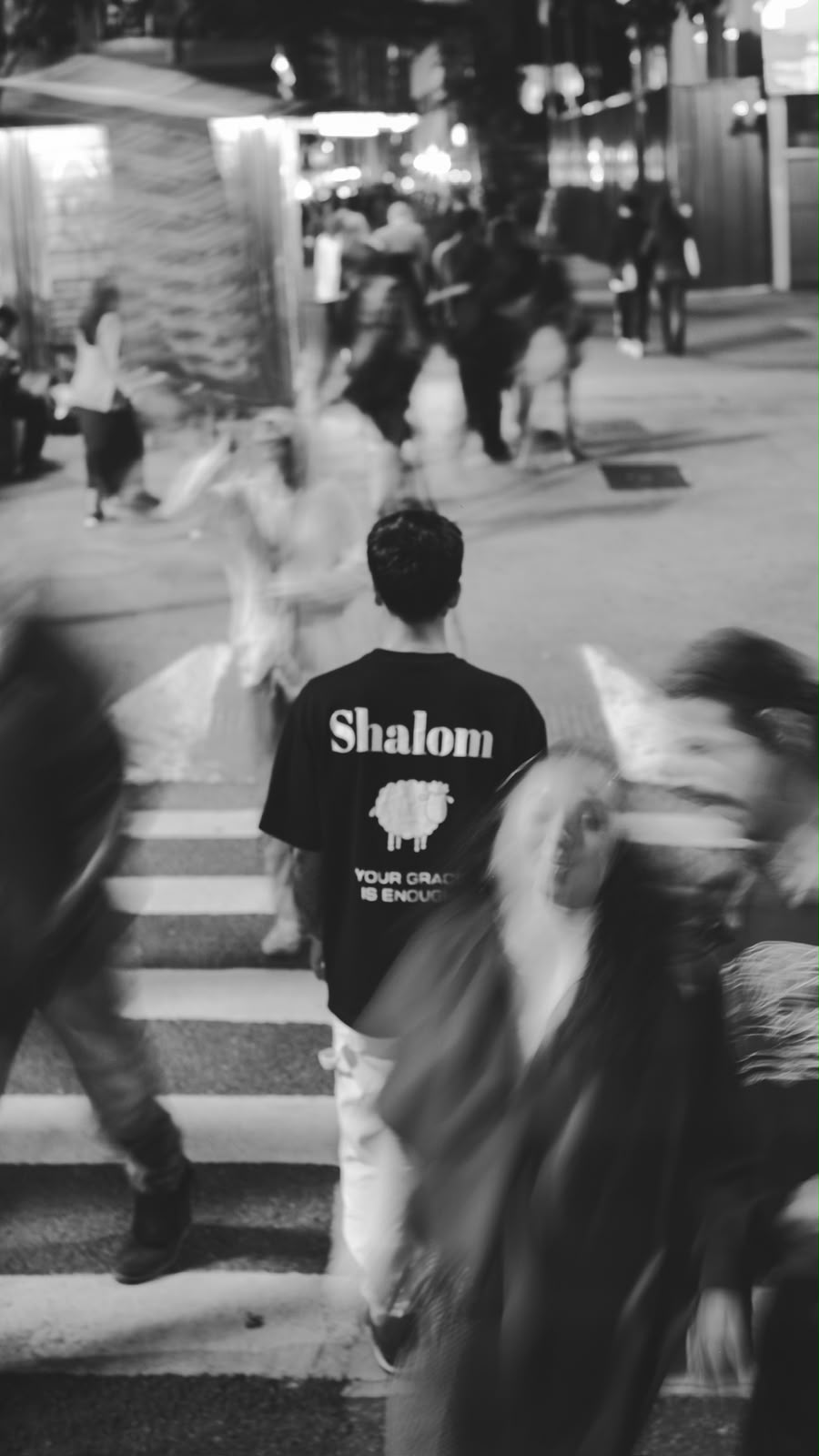 Jovem atravessando faixa de pedestres com camiseta preta SHALOM Your Grace Is Enough — foto preto e branco