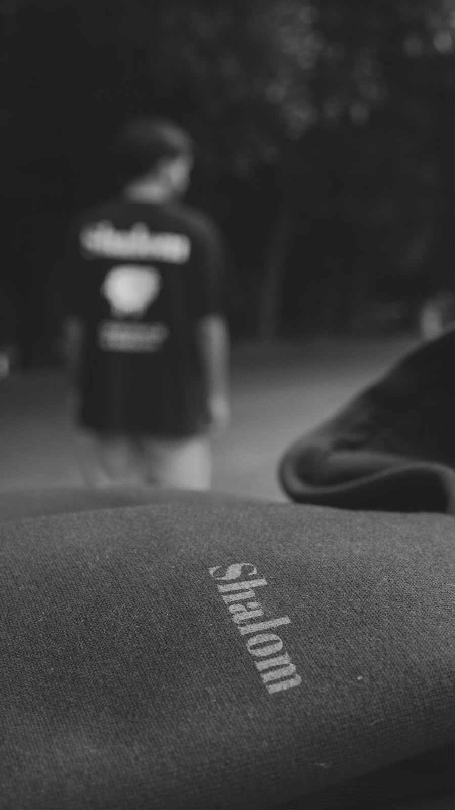 Detalhe de camiseta SHALOM com pessoa desfocada ao fundo usando camiseta preta SHALOM — foto preto e branco